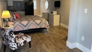 A bedroom with a bed, a chair, a desk, a TV, and a kitchen area in the background. - Bavarian Brook Unit 8 (Helen)
