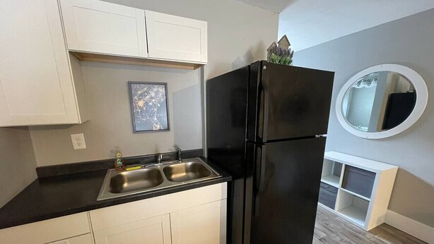A compact kitchen with white cabinets, a black refrigerator, a double sink, and a small round mirror on the wall. - Bavarian Brook Unit 8 (Helen)