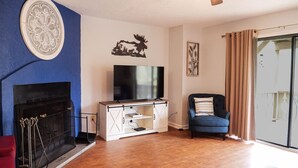 Un salon avec une cheminée, un téléviseur posé sur un meuble blanc, un fauteuil bleu et un ventilateur de plafond.