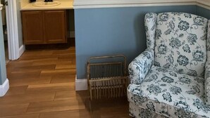 A room with a wooden floor, a blue accent wall, a white chair with a floral pattern, a wooden bench, a painting of a forest path, and a bathroom with a sink and mirror. - Rivers Edge 802 (Helen)