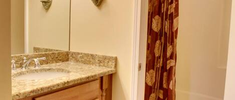 A bathroom with a granite countertop sink, wooden cabinets, a toilet, and a shower curtain with a leaf pattern.