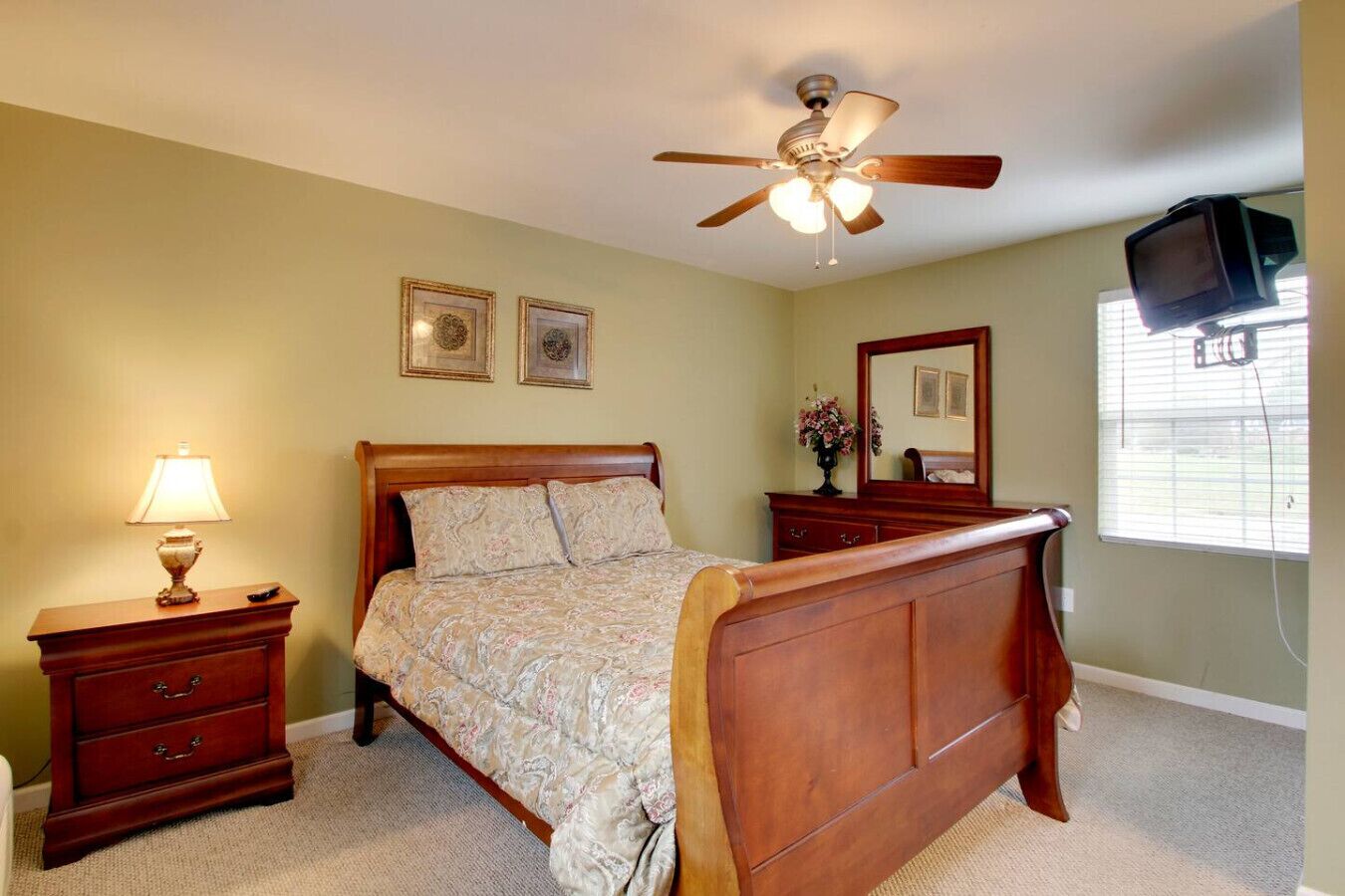 A bedroom with a wooden bed, a nightstand with a lamp, a ceiling fan, a dresser, a mirror, and a television mounted on the wall.