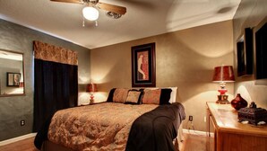 Une chambre avec un grand lit, un ventilateur de plafond, une table de chevet avec une lampe et un tableau encadré au mur.