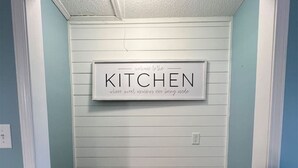 Un corridoio stretto con un tavolo e panche di legno, una insegna che riporta la scritta “WELCOME TO THE KITCHEN” e pareti azzurro chiaro.