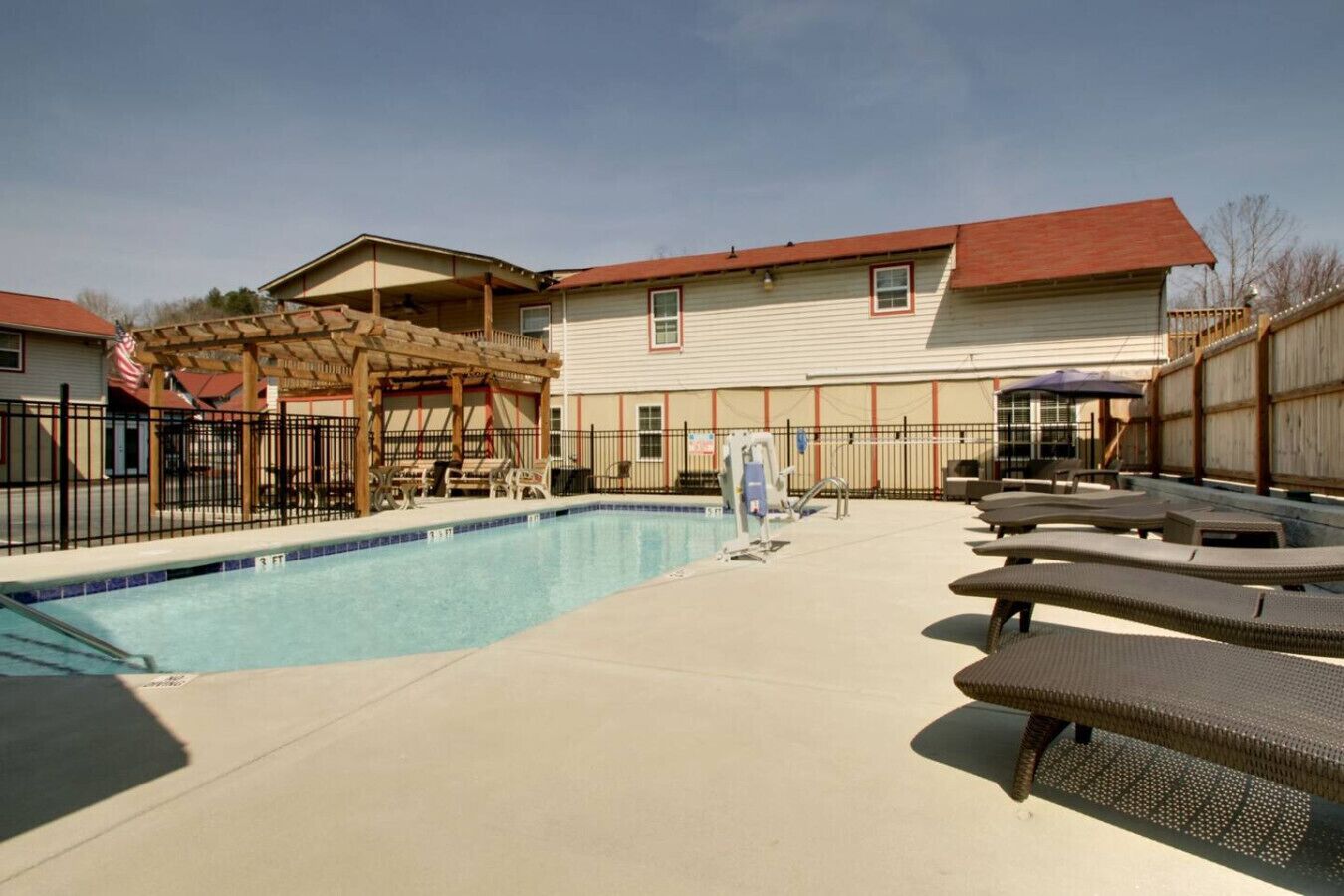 A swimming pool with lounge chairs, a covered seating area, and a building with a red roof in the background.