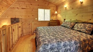 A cozy wooden cabin bedroom with a bed, a nightstand, a dresser, a window, and a wooden railing. - Bavarian Hideaway 24 (Sautee Nacoochee)