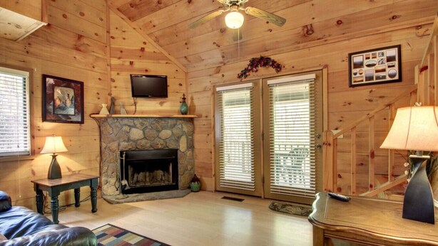 A cozy log cabin interior with a stone fireplace, a ceiling fan, and a television mounted on the wall. - Bavarian Hideaway 24 (Sautee Nacoochee)