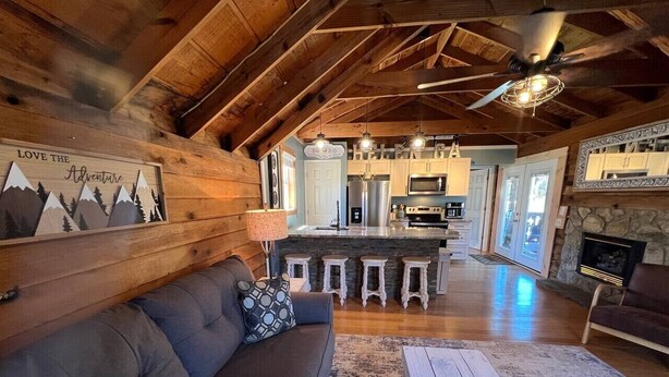 A cozy cabin interior with a wooden ceiling, a living area with a sofa and a fireplace, a kitchen with bar stools, and a stone fireplace. - Bavarian Brook Condo 14 (Helen)