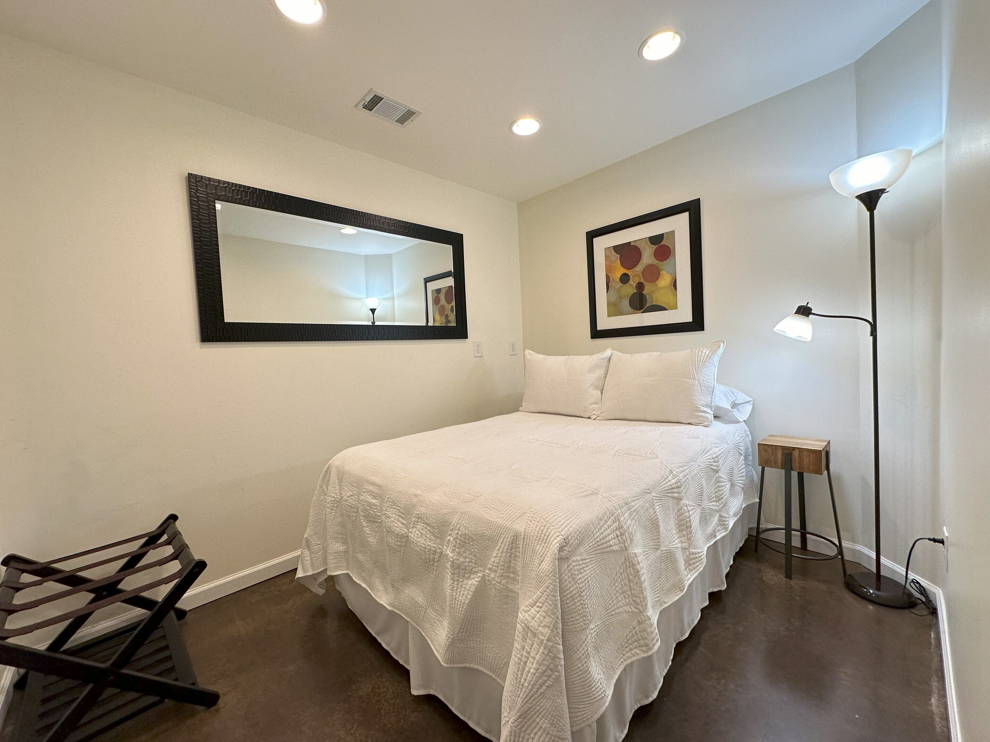 A neatly arranged bedroom with a bed, a mirror, a painting, and a floor lamp.