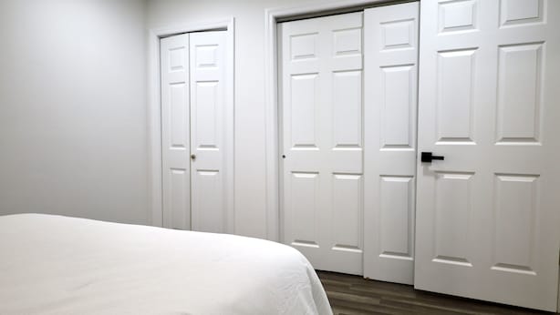 Ein Schlafzimmer mit einem Bett, weißen Wänden und Holzboden. Zwei weiße Türen und ein Kleiderschrank sind ebenfalls vorhanden.
