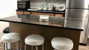 Eine Küche mit Holzausstattung, einem Edelstahlkühlschrank, einer Mikrowelle und einer Kaffeemaschine. Drei weiße Barhocker mit Metallbeinen sind ebenfalls vorhanden.