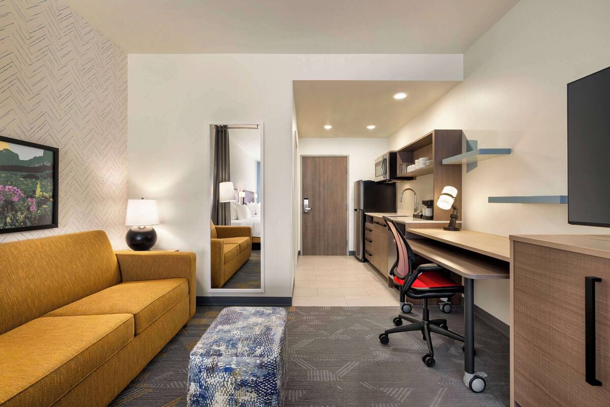 A hotel room with a sofa, a desk with a chair, a mirror, a television, and a kitchenette.