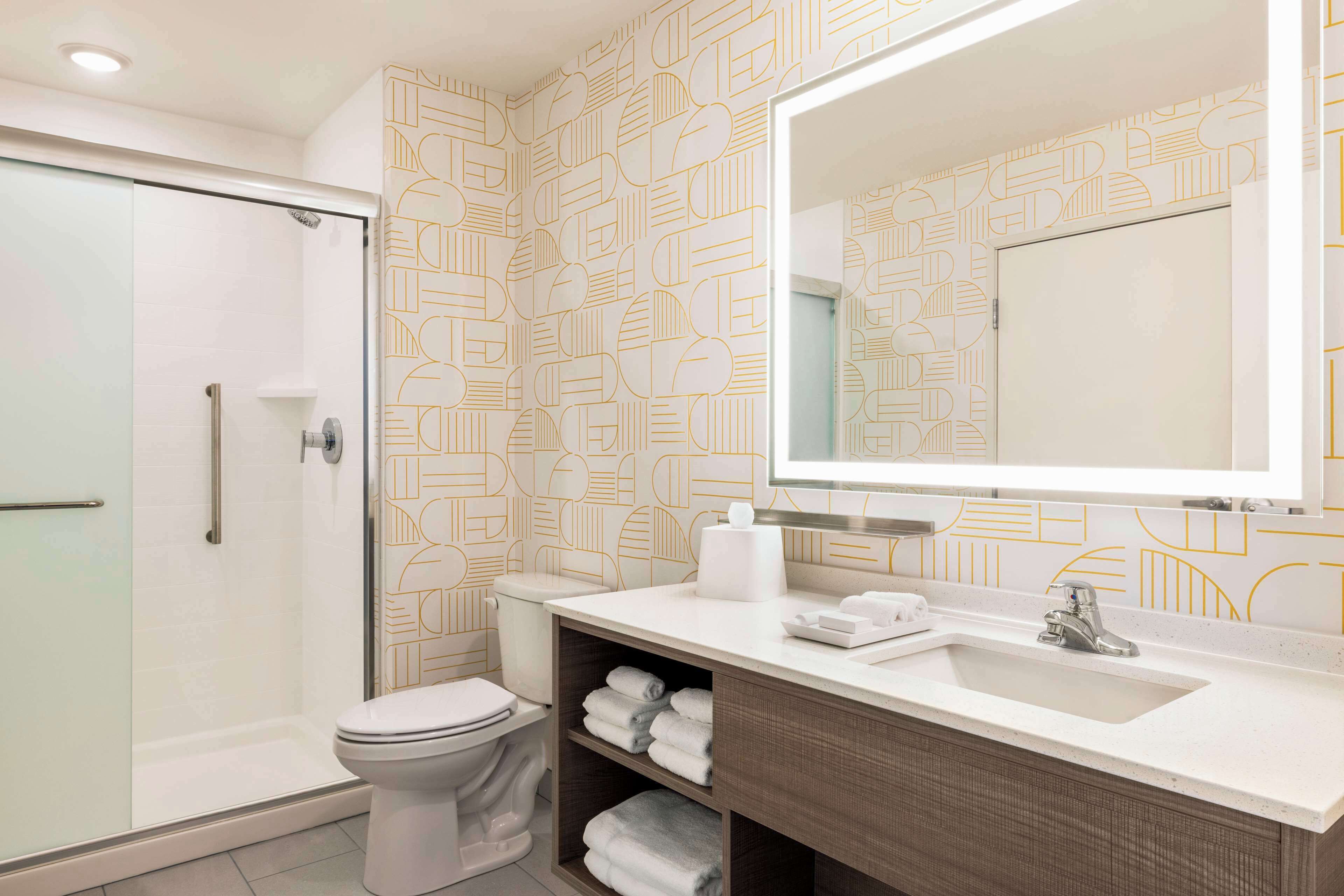 A modern bathroom with a glass shower enclosure, a toilet, a vanity with a large mirror, and shelves with folded towels.