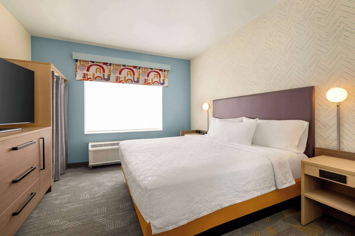 A hotel room with a bed, a desk, a dresser, a window with curtains, and a wall with a geometric pattern.