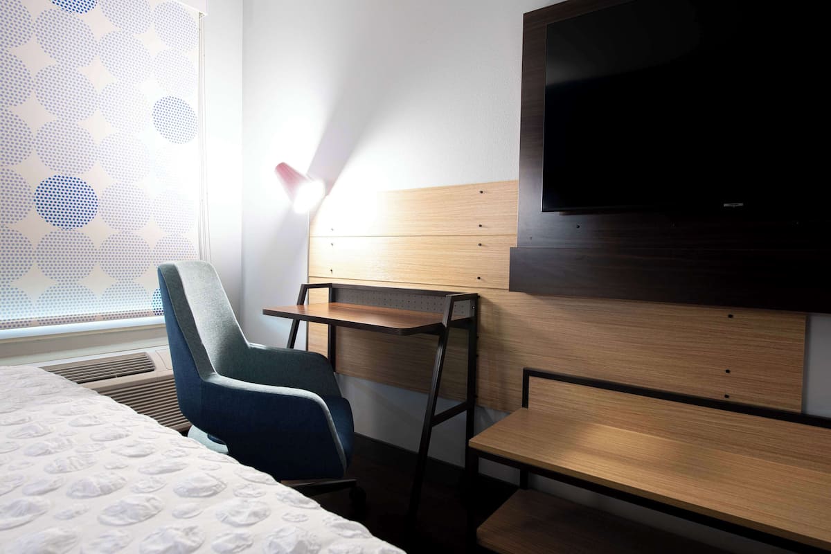 A hotel room with a bed, a desk, a chair, a TV, and a window with a geometric pattern.