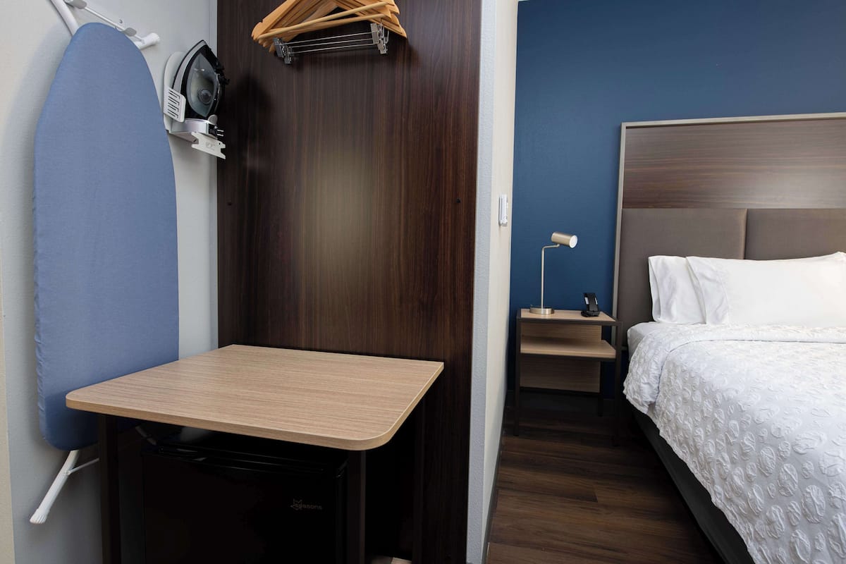 A compact hotel room with a wooden table, a bed with white bedding, a nightstand with a lamp, and a wall-mounted ironing board.