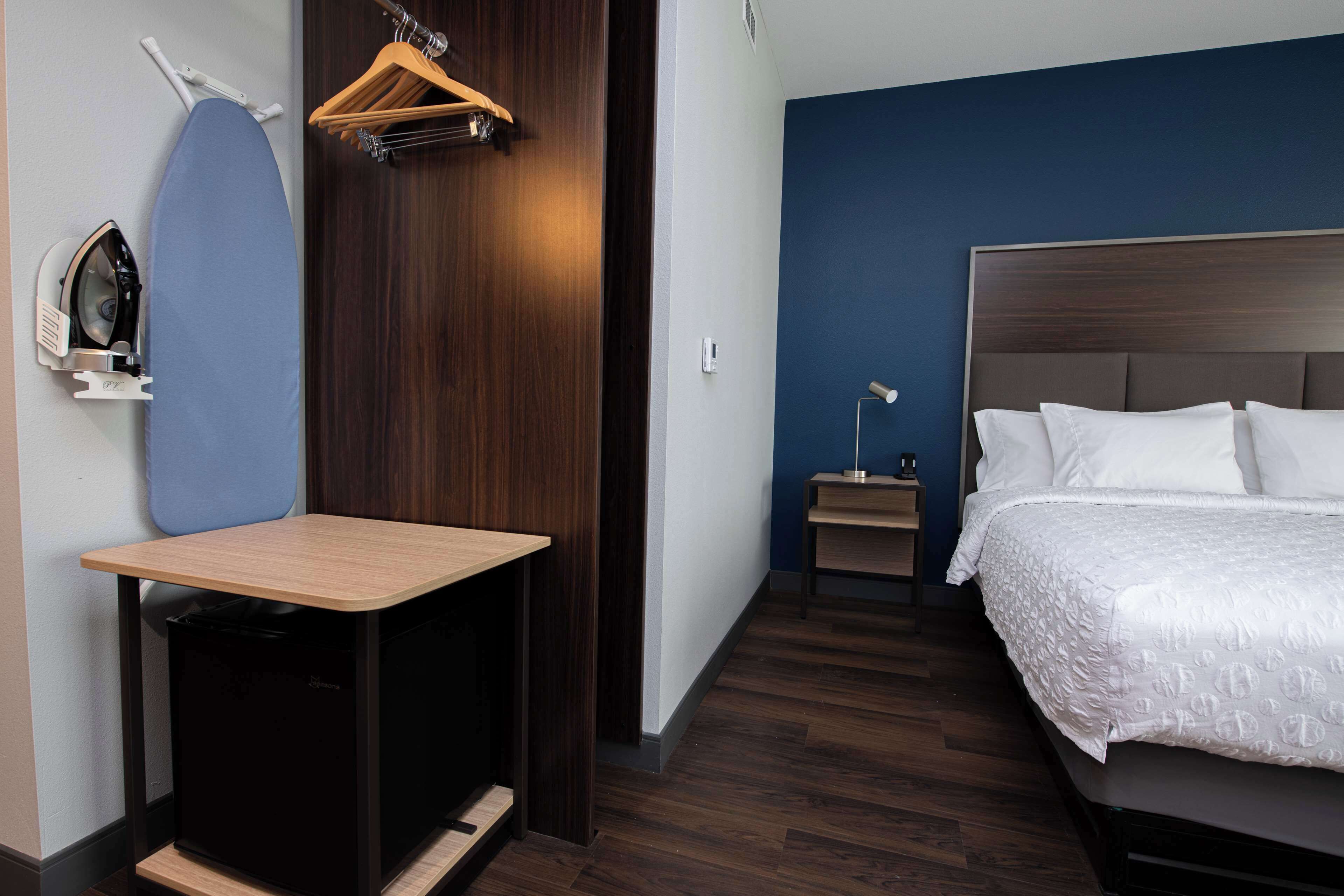Una habitación de hotel con cama, escritorio con tabla de planchar, mesita de noche pequeña y armario.
