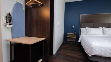 Una habitación de hotel con cama, escritorio con tabla de planchar, mesita de noche pequeña y armario.