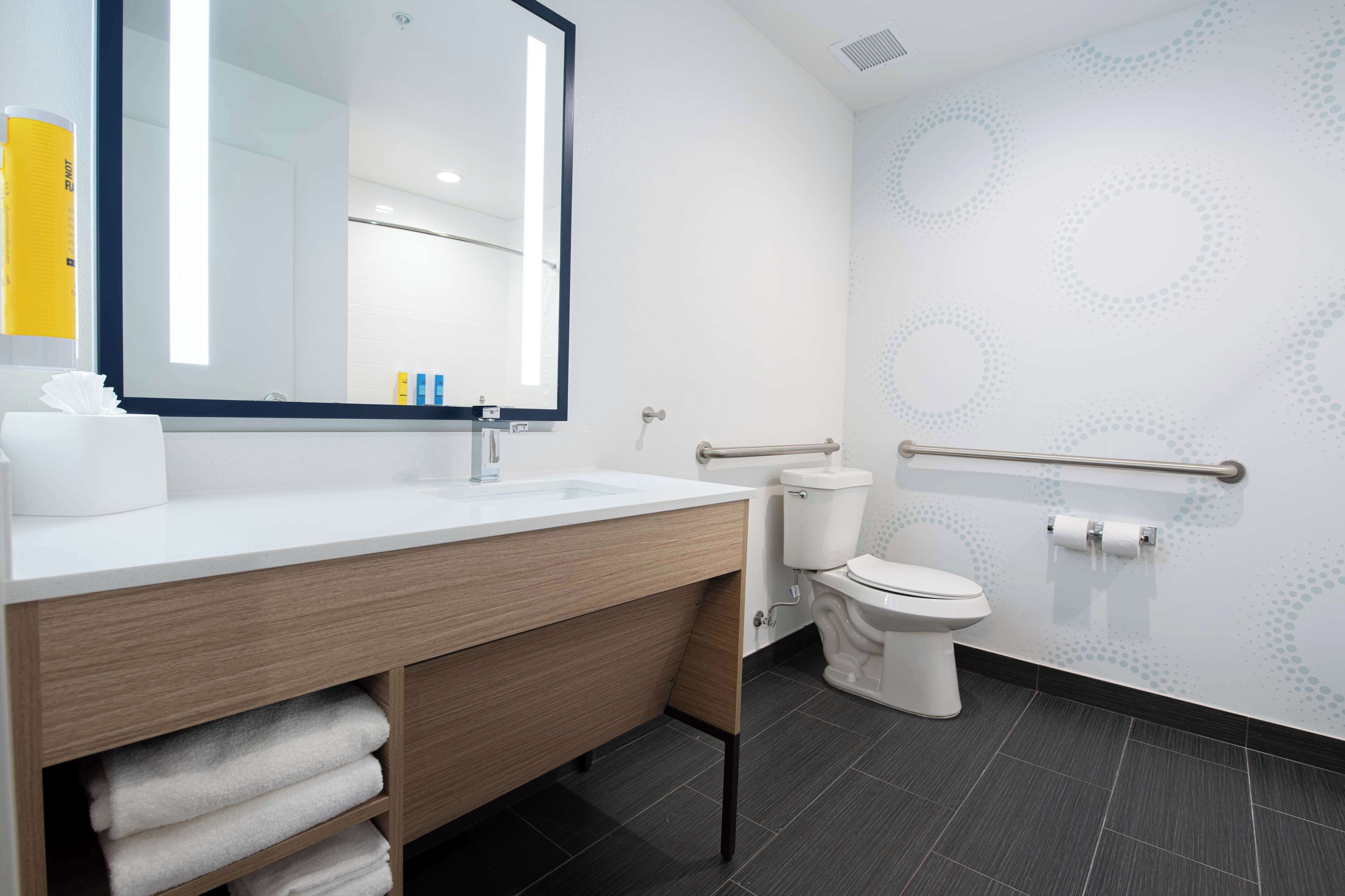 Un baño moderno con lavamanos, espejo y retrete blancos. Hay toallas en el mostrador y un pasamanos cerca del retrete.