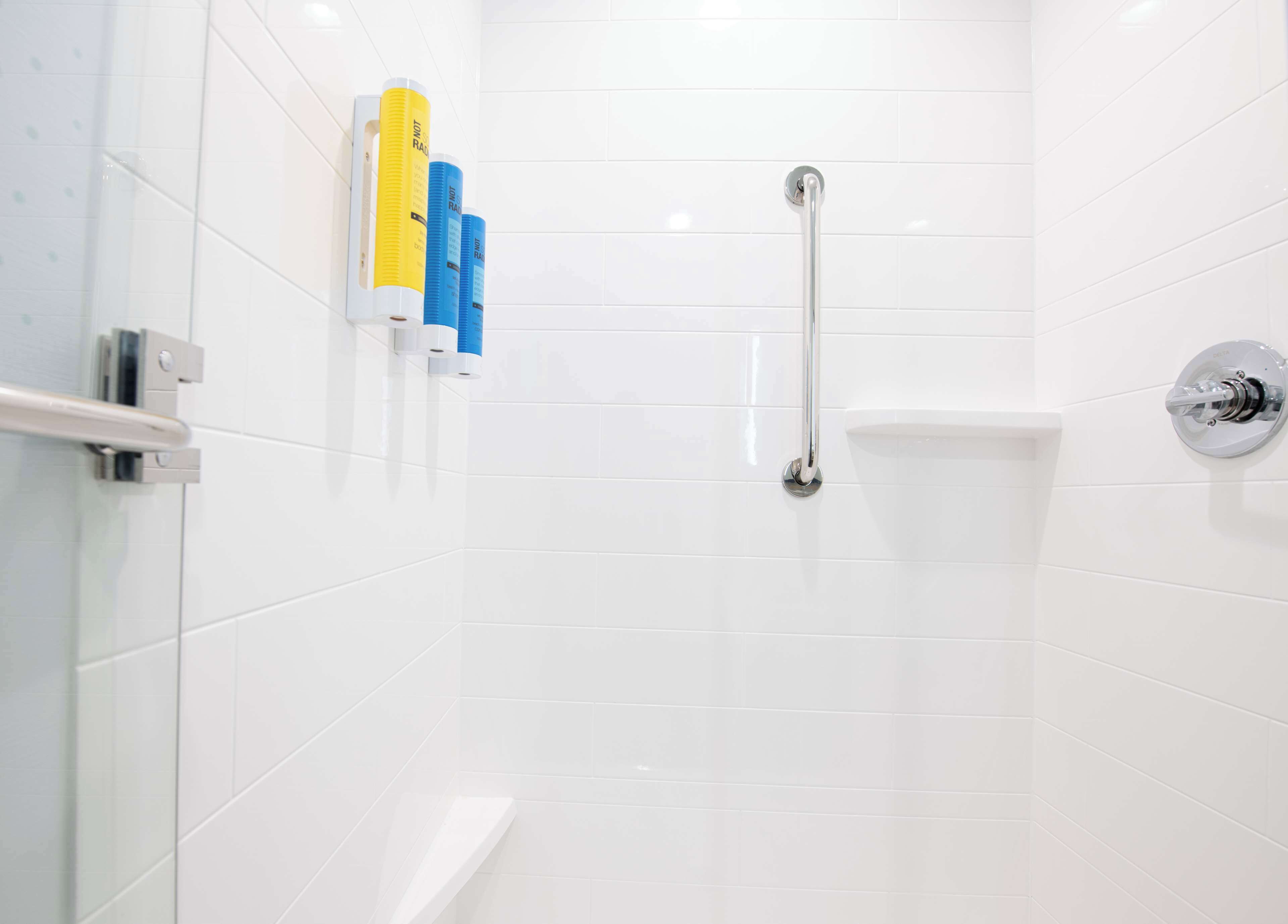 Une salle de bain moderne avec des carreaux blancs, une cabine de douche en verre, une douchette à main et un distributeur de papier toilette.