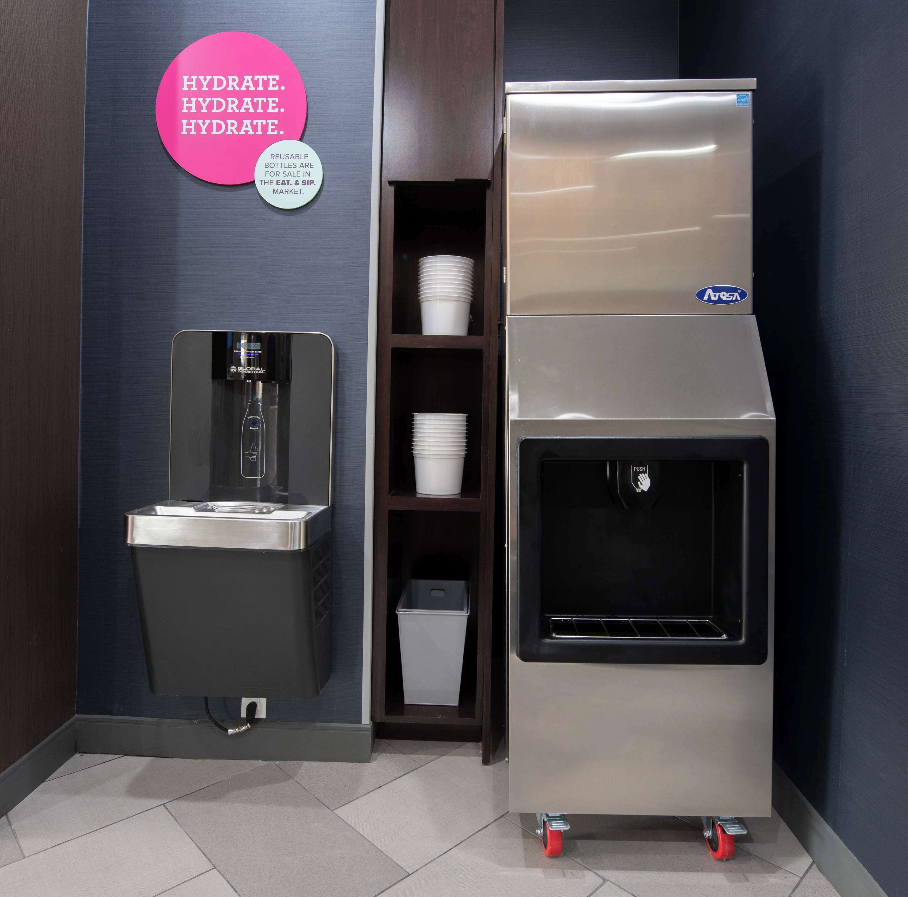 A water dispenser with a push button, a sink, and a shelf with white cups and a trash bin.