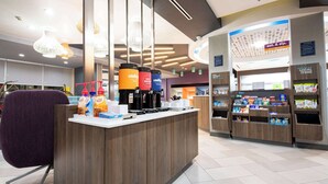 A modern coffee shop with a wooden counter, coffee machines, and a variety of beverages. There are hanging lights and a sign for local items. - Tru By Hilton Lewisville Vista Ridge Mall (Lewisville)