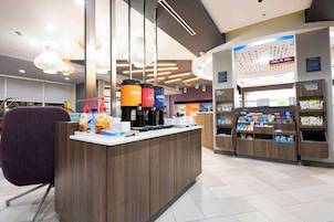 A modern coffee shop with a wooden counter, coffee machines, and a variety of beverages. There are hanging lights and a sign for local items.