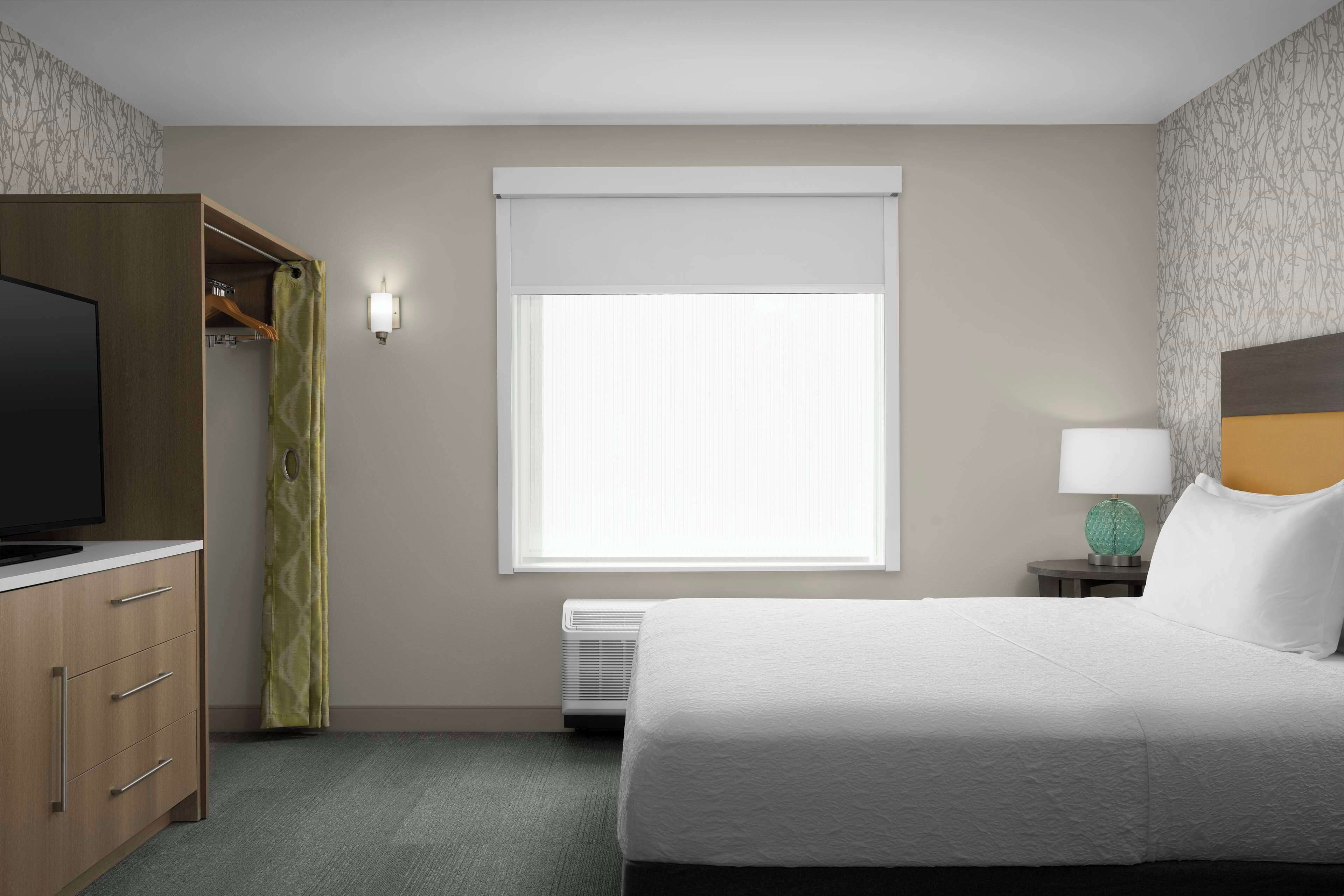 A hotel room with a bed, a TV, a wardrobe, a window with blinds, a lamp, and an air conditioner.