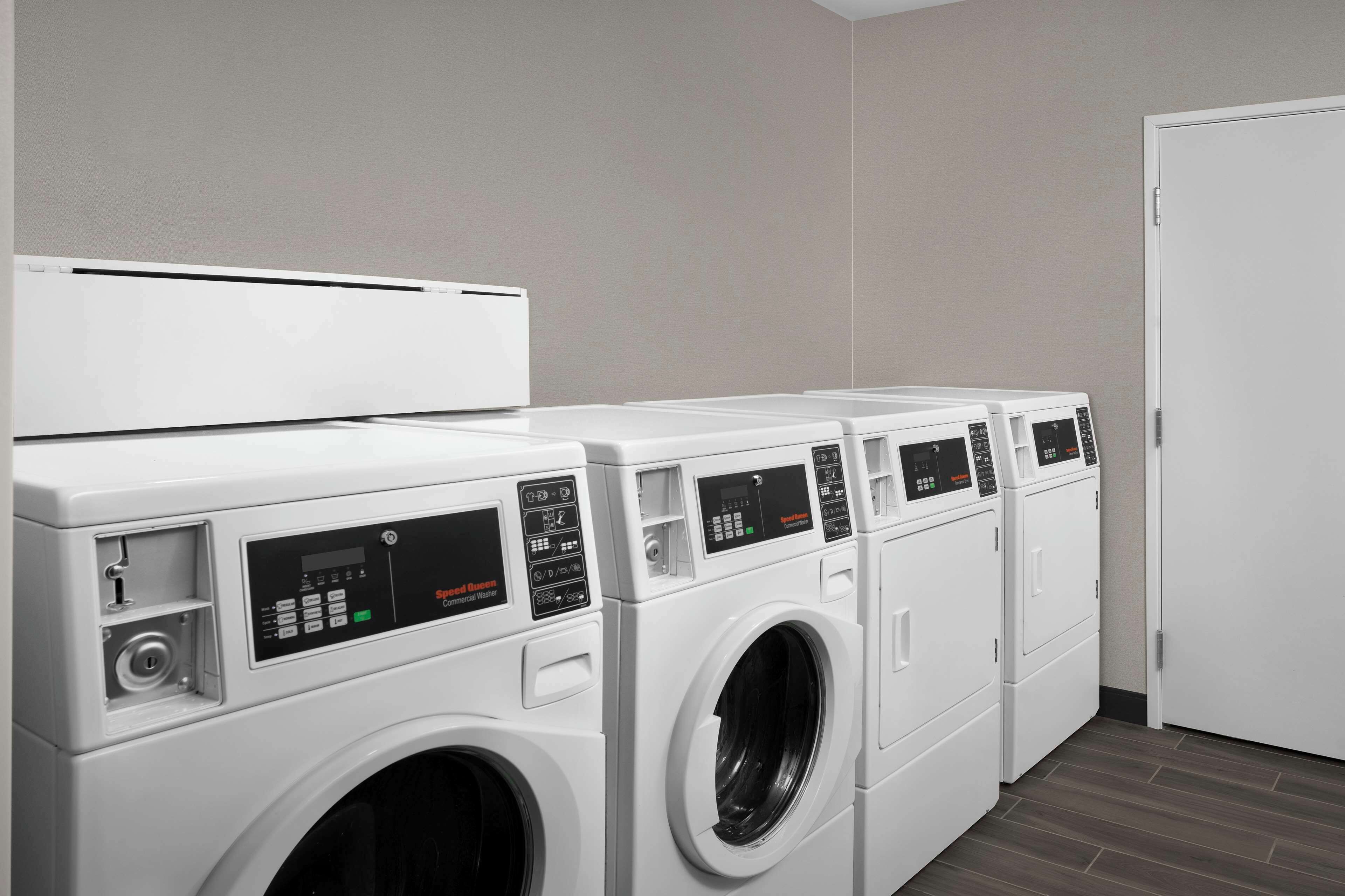 A row of white front-loading washers in a laundry room with a plain wall and a door on the right.