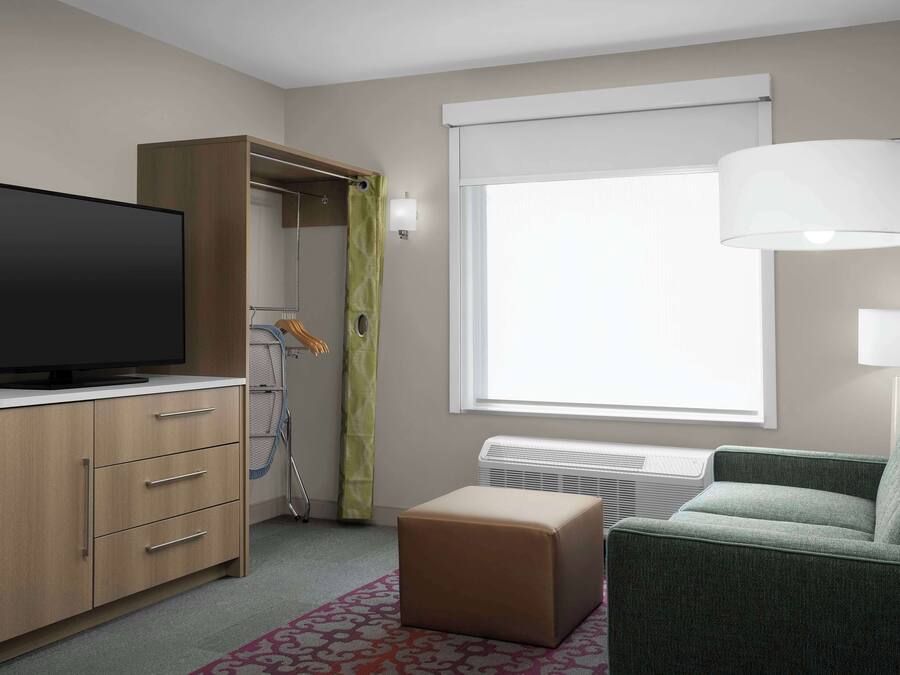 Ein Hotelzimmer mit Flachbildfernseher, einem Holzschrank, einem grünen Sofa, einem gemusterten Teppich und einem großen Fenster mit weißem Vorhang.