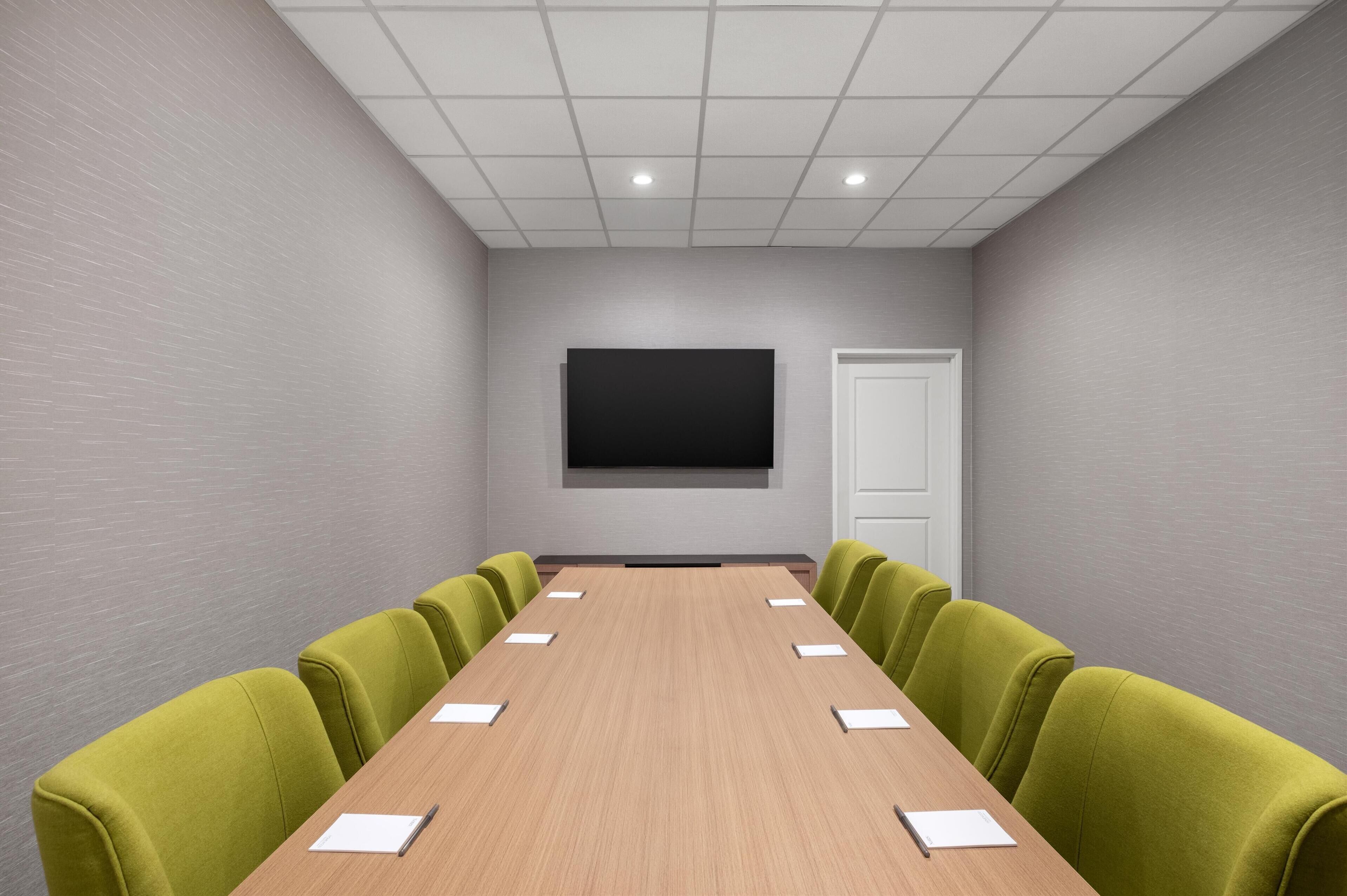 Una sala de conferencias con una mesa larga, sillas verdes, una pantalla grande y una puerta.