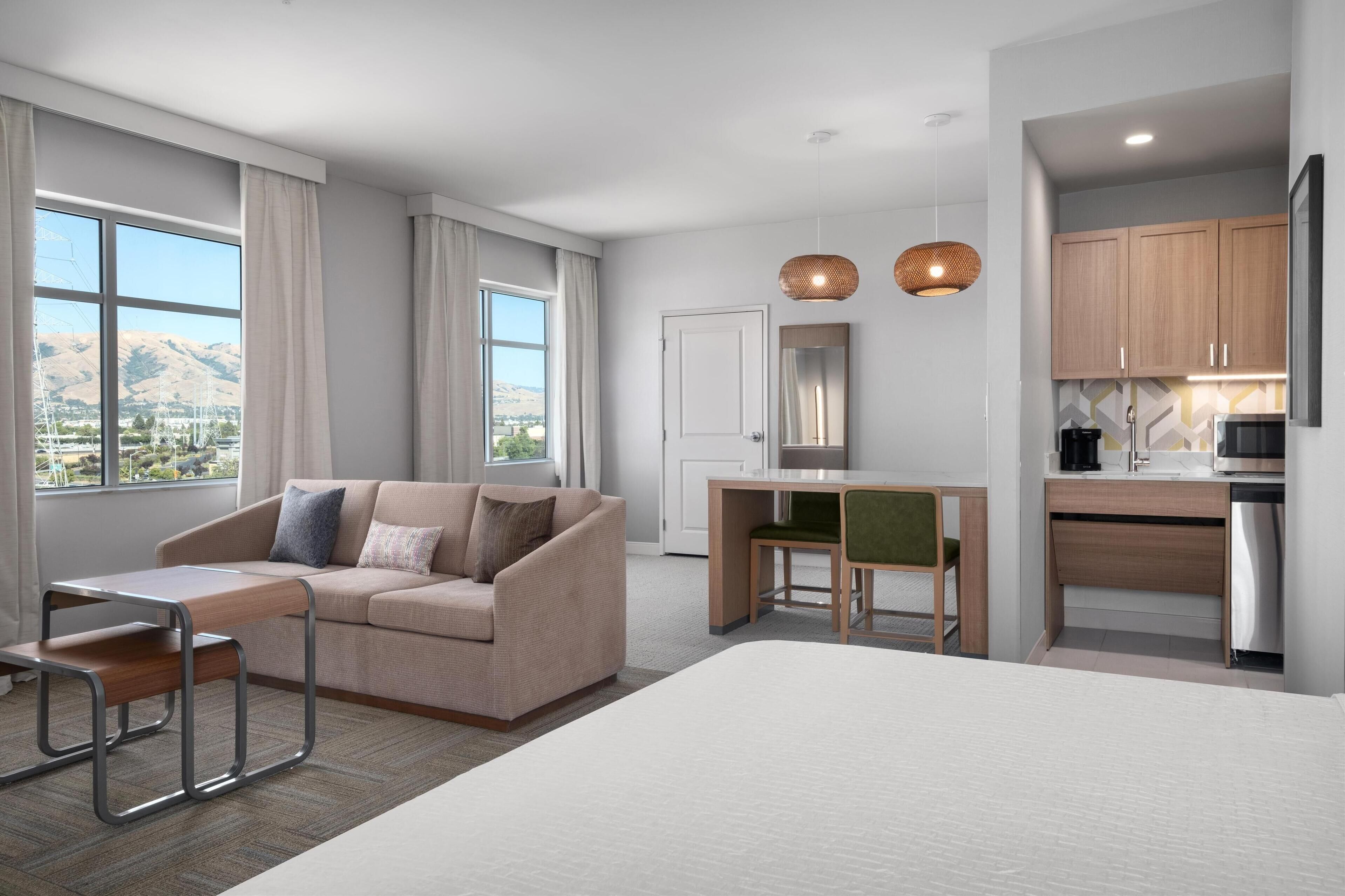 A modern hotel room with a sofa, a desk, and a bed. There is a kitchen area with cabinets and a refrigerator.