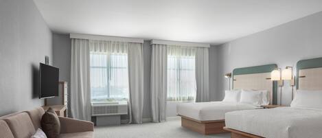 Une chambre d’hôtel avec deux lits, un canapé, un téléviseur à écran plat et de grandes fenêtres équipées de rideaux.
