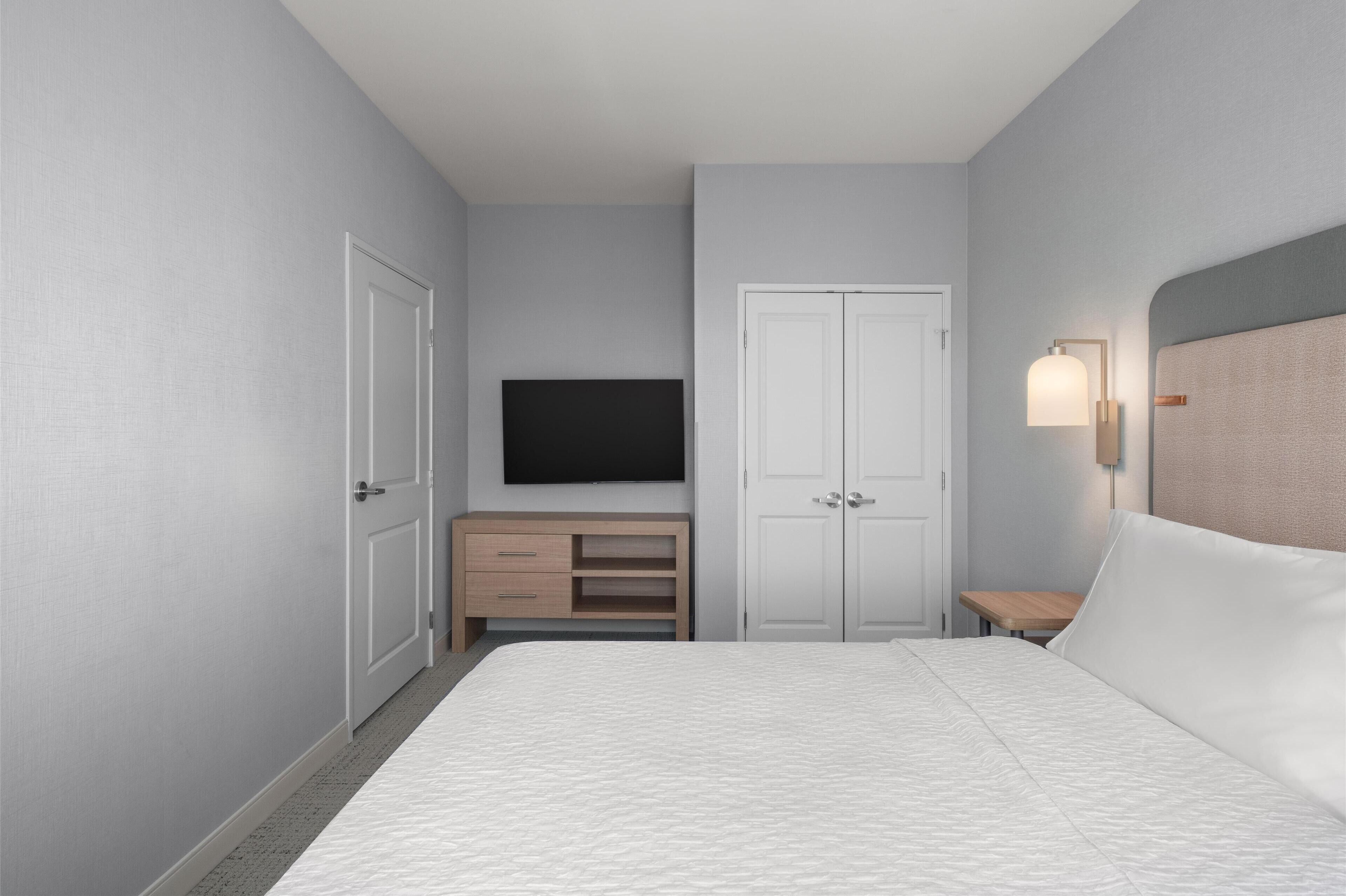 A bedroom with a bed, a TV mounted on the wall, a wardrobe, a bedside table, and a lamp.