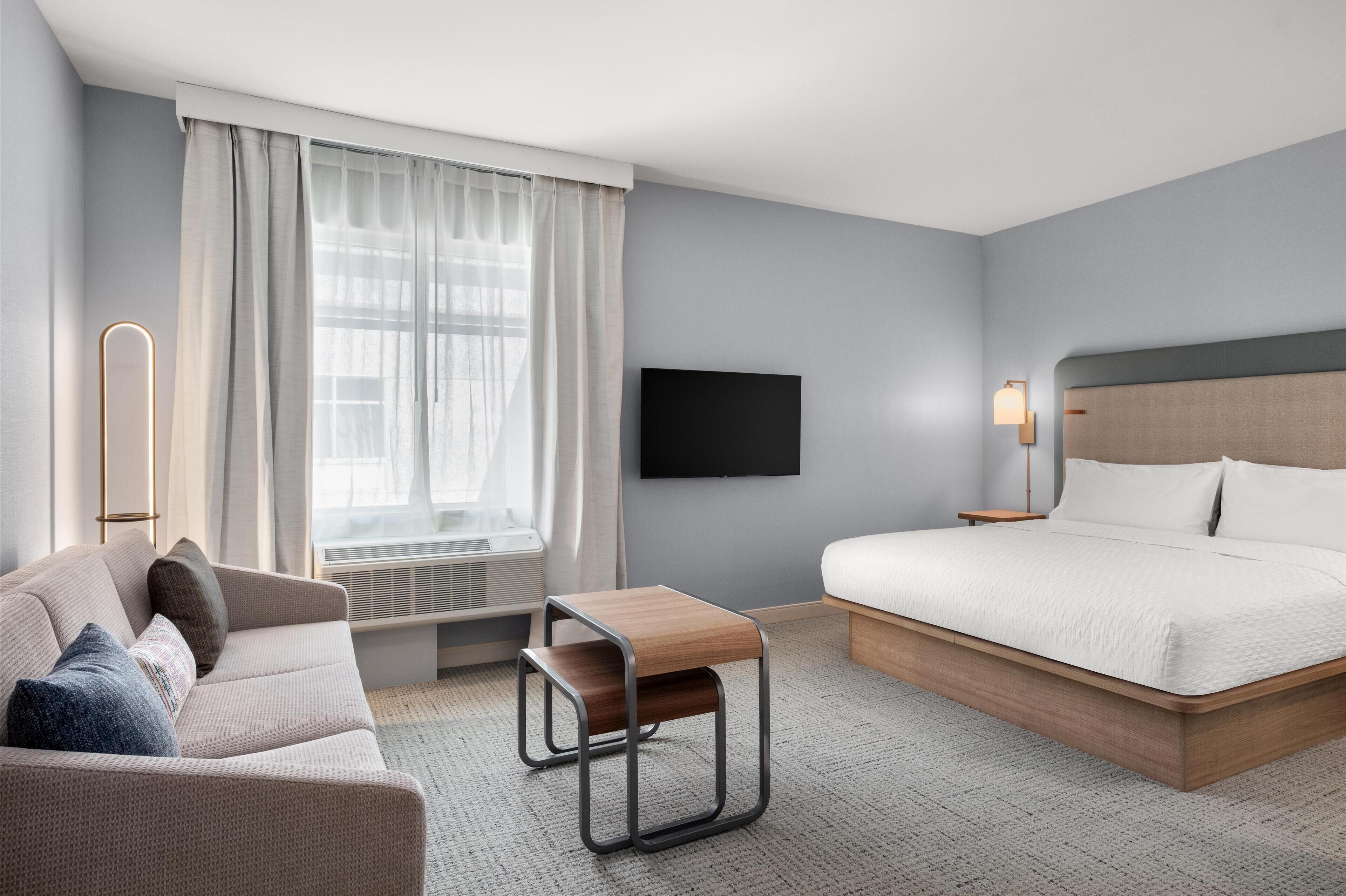 A hotel room with a bed, a sofa, a small wooden table, a TV mounted on the wall, and a window with curtains.