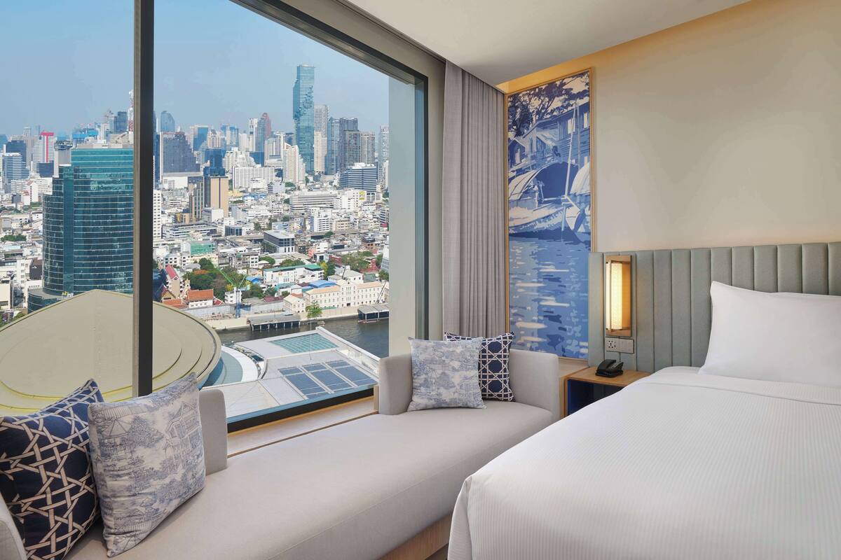 A hotel room with a large window offering a city view, a bed, a seating area with a sofa and pillows, and a small table with a phone.