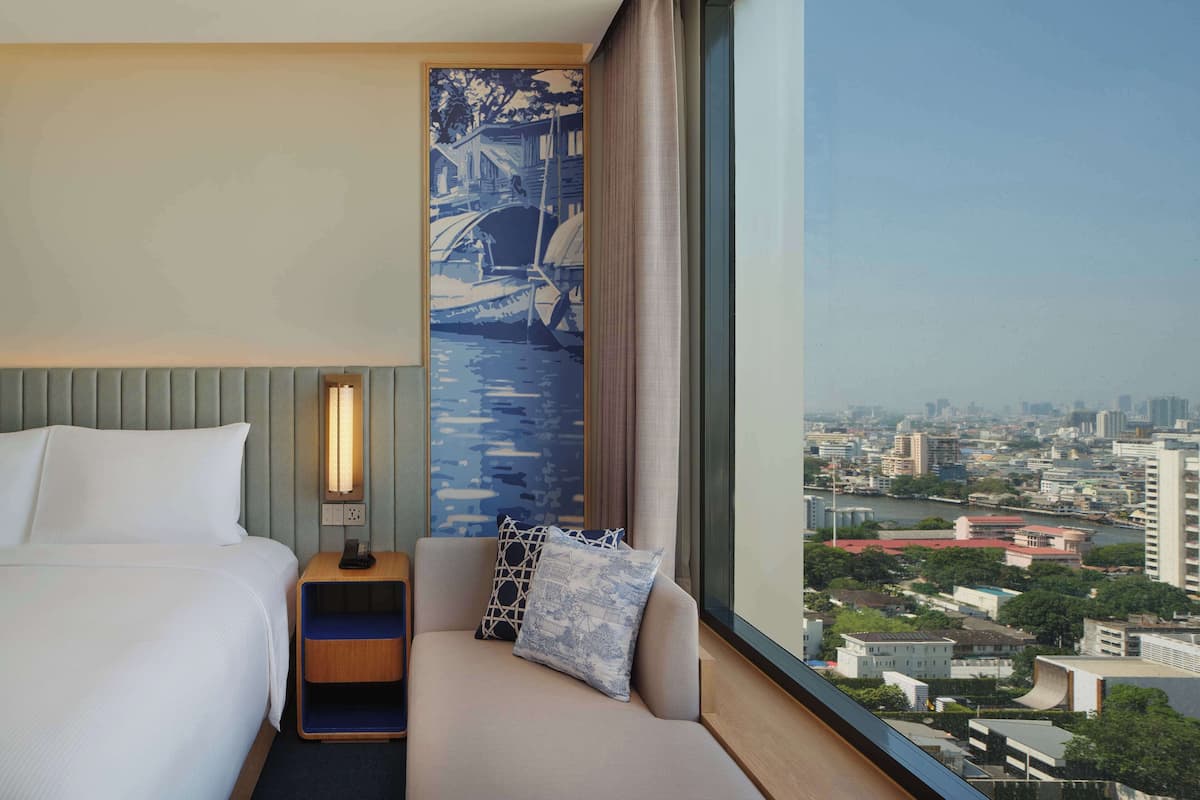 A hotel room with a bed, a bedside table with a lamp, a sofa with cushions, and a large window offering a city view.