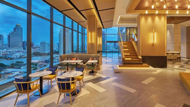 A modern interior with large windows, a staircase, and a dining area with tables and chairs. - Hilton Garden Inn Bangkok Riverside (Bangkok)