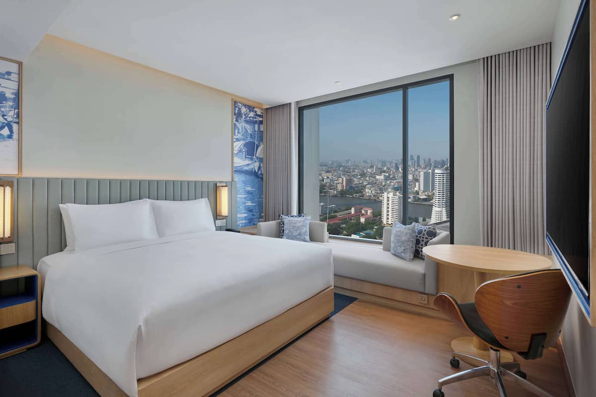 A modern hotel room with a large bed, a desk, and a view of a cityscape.