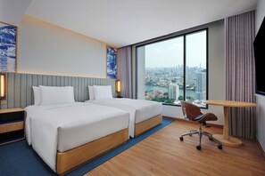 Habitación de hotel con dos camas, un escritorio, una silla, un ventanal grande con vista a la ciudad y un televisor de pared.