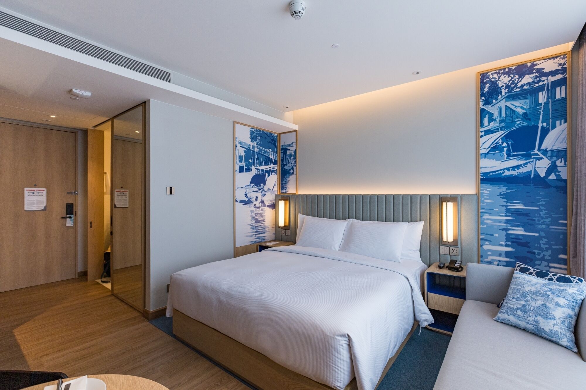 A hotel room with a large bed, a smaller bed, a desk, and a chair. There are two wall-mounted lamps and a ceiling-mounted security camera.
