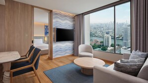 A modern hotel room with a large window, a flat-screen TV, a dining area, and a sofa. - Hilton Garden Inn Bangkok Riverside (Bangkok)