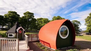 A red and green geodesic dome structure with a small adjacent wooden cabin, both situated in a grassy area with trees in the background.