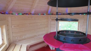A wooden cabin interior with a round grill table, wooden benches, and a window with a view of greenery.