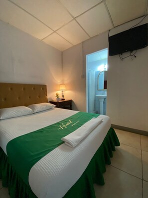 A hotel room with a bed, a nightstand, a lamp, a television, and a bathroom visible through an open door. - Hostal Pochote  (León)