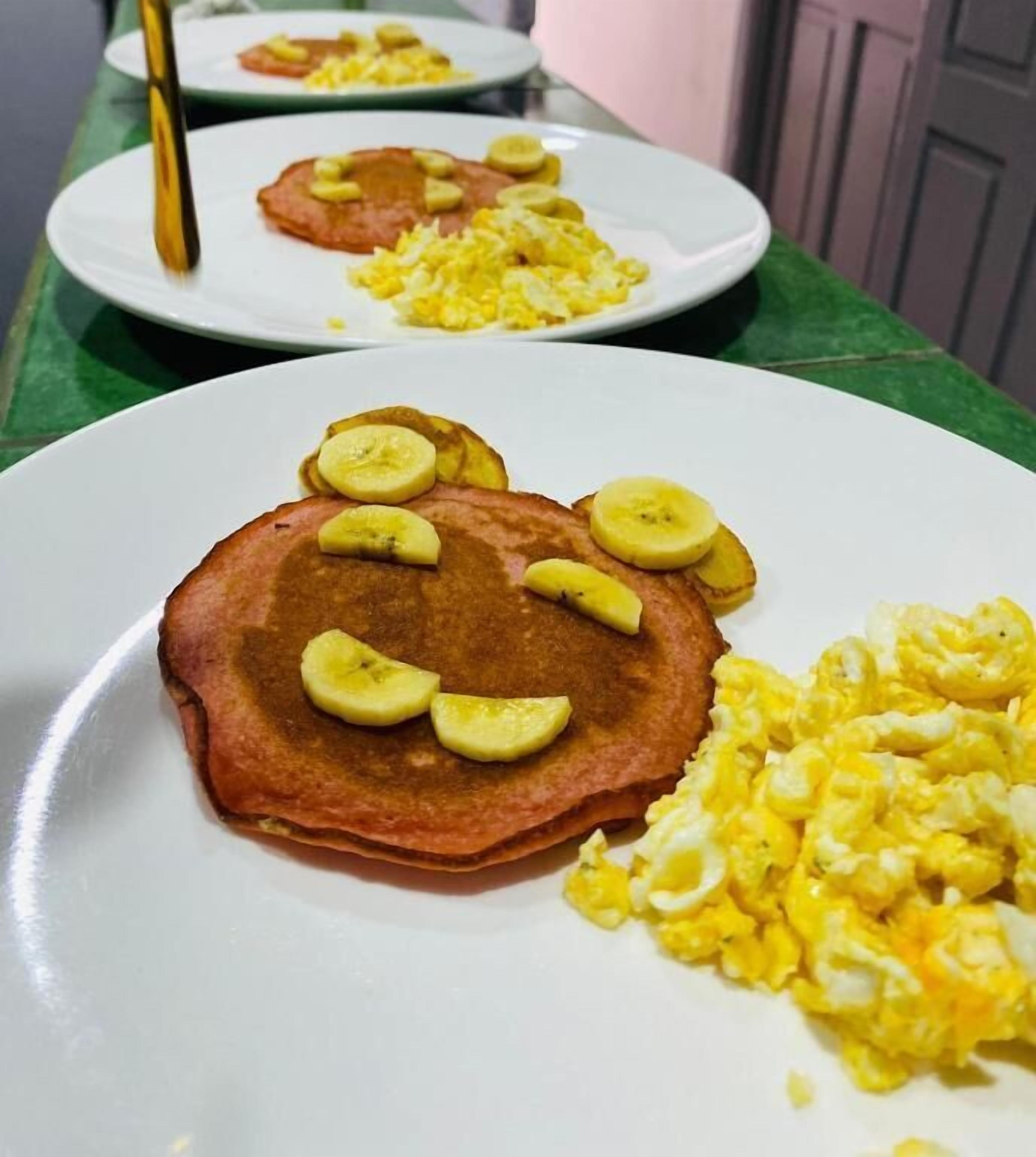 Desayuno con tortitas cubiertas de rodajas de plátano, huevos revueltos y una cuchara.