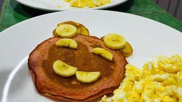 Desayuno con tortitas cubiertas de rodajas de plátano, huevos revueltos y una cuchara.
