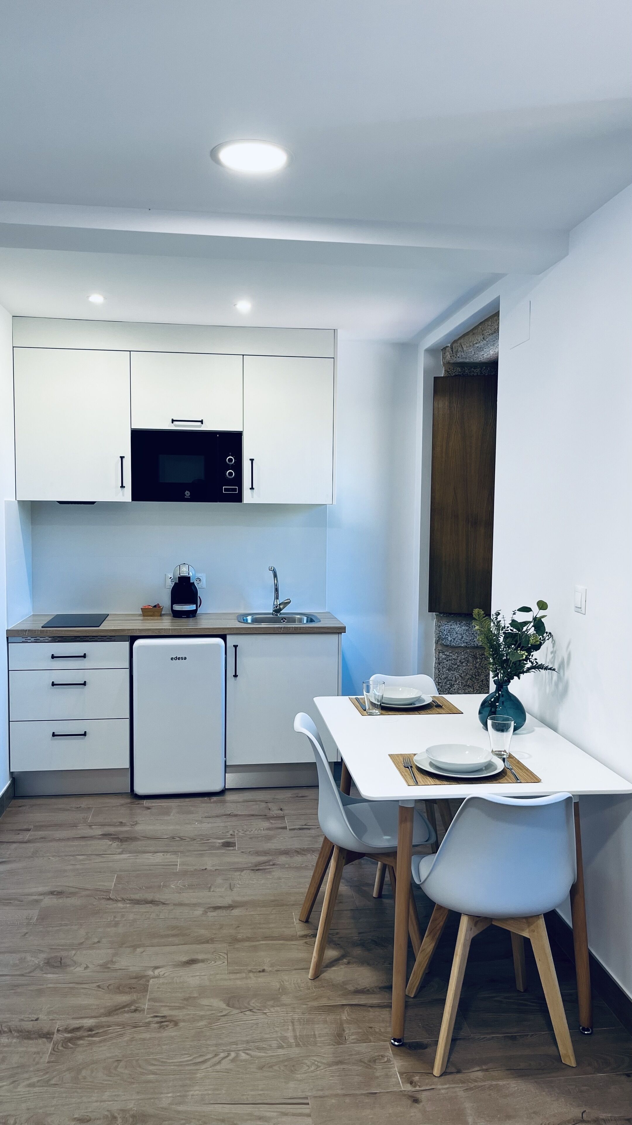 Eine moderne Küche mit weißen Schränken, einer Mikrowelle und einem Spülbecken. Ein Esstisch mit Stühlen und eine Pflanze an der Wand sind ebenfalls vorhanden.