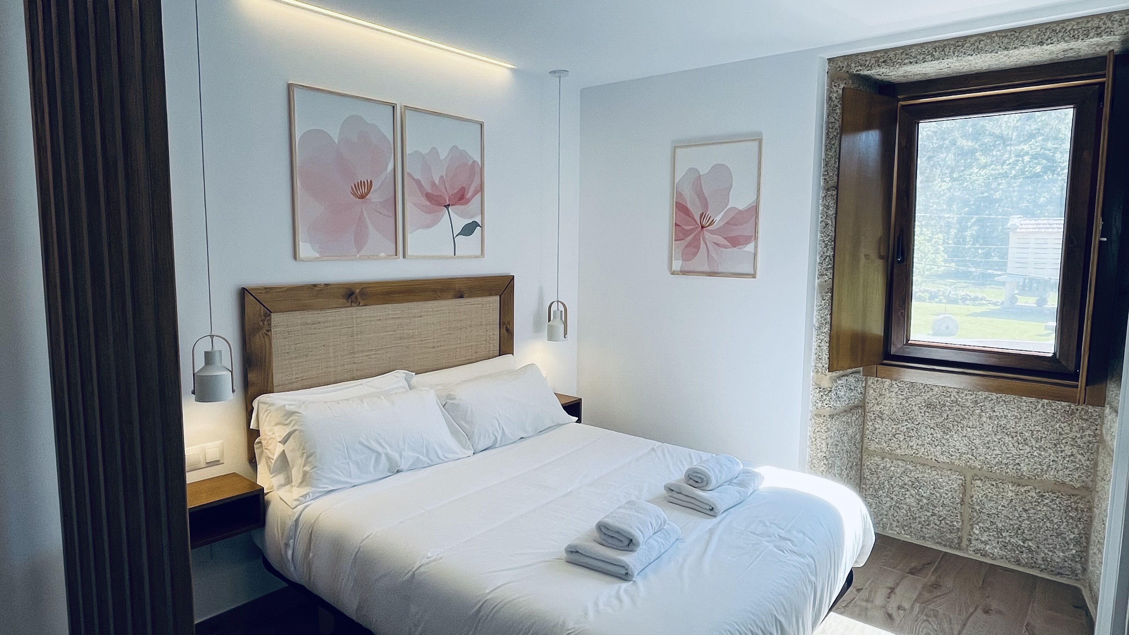 Una cama bien tendida con sábanas blancas y dos toallas enrolladas, un cabecero de madera y una mesita de noche con una lámpara.