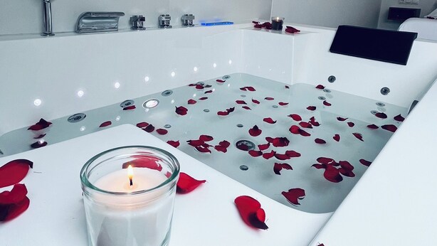 Un baño moderno con bañera, una vela de cristal y pétalos de rosa en el suelo.
